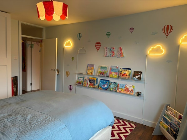 A bedroom with a bed, a wall-mounted bookshelf with children's books, and a door leading to another room.