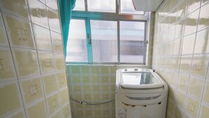 A small, narrow bathroom with yellow patterned tiles, a washing machine, and a window with a curtain. - Apto com Wi-fi Ar-condicionado e Vaga de Garagem (Guarujá)