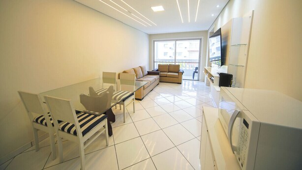 A modern living room with a glass dining table, a sofa, and a TV. - Apto com Wi-fi Ar-condicionado e Vaga de Garagem (Guarujá)