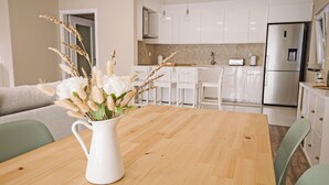 A modern kitchen with white cabinets, a stainless steel refrigerator, and a wooden dining table with a white pitcher of flowers. - Close to Lisbon & Beaches (Amora)