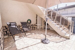 An outdoor patio area with a stone floor, a covered seating area with chairs, a table, and a staircase leading to an upper level.