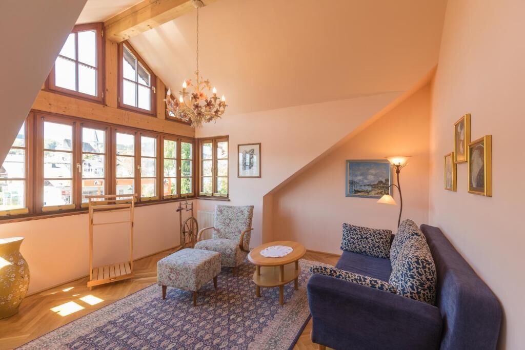 Ein gemütliches Loft-Zimmer mit hoher Decke, großen Fenstern, einer Kristallleuchter, einer Couch, einem Sessel und einem kleinen runden Tisch.