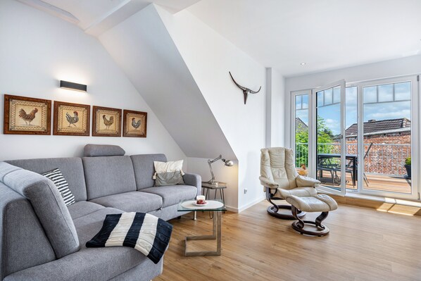 Un salon moderne avec un canapé gris, une table basse en verre et un fauteuil en cuir. Des tableaux encadrés ornent le mur, ainsi qu’une décoration représentant une tête de taureau. De grandes fenêtres offrent une vue sur un bâtiment en briques.