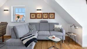 Una sala de estar moderna con un sofá seccional gris, una mesa de centro de vidrio, una mesita auxiliar con lámpara y una ventana que ofrece vistas a una casa.