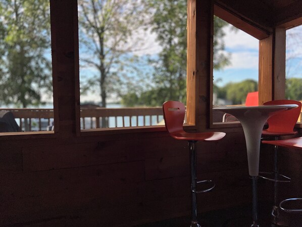 Un intérieur en bois avec deux tabourets de bar et une table, donnant sur une étendue d’eau bordée d’arbres et d’un quai.