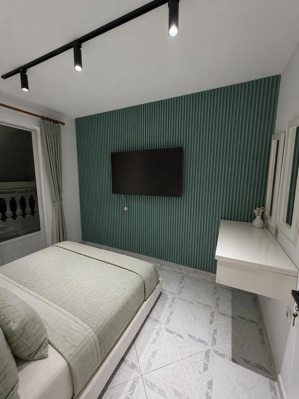 Ein Schlafzimmer mit Bett, einem an der Wand befestigten Fernseher, einem Nachttisch und einem Fenster mit Vorhängen.