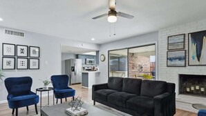 Una sala de estar moderna con un sofá de cuero negro, dos sillones azules y una mesa de centro. Hay una chimenea con pared de ladrillo y un ventilador de techo.