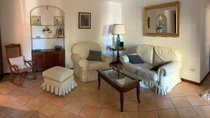 Un salon comprenant un canapé, un fauteuil, un pouf et une table basse. On y trouve une lampe sur une table d’appoint, un miroir au mur et une étagère avec des objets décoratifs.