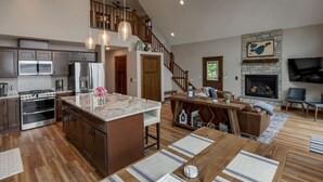 Amplia sala de estar con un gran comedor de madera, una cocina equipada con electrodomésticos modernos, una chimenea y una escalera que conduce a un nivel superior.