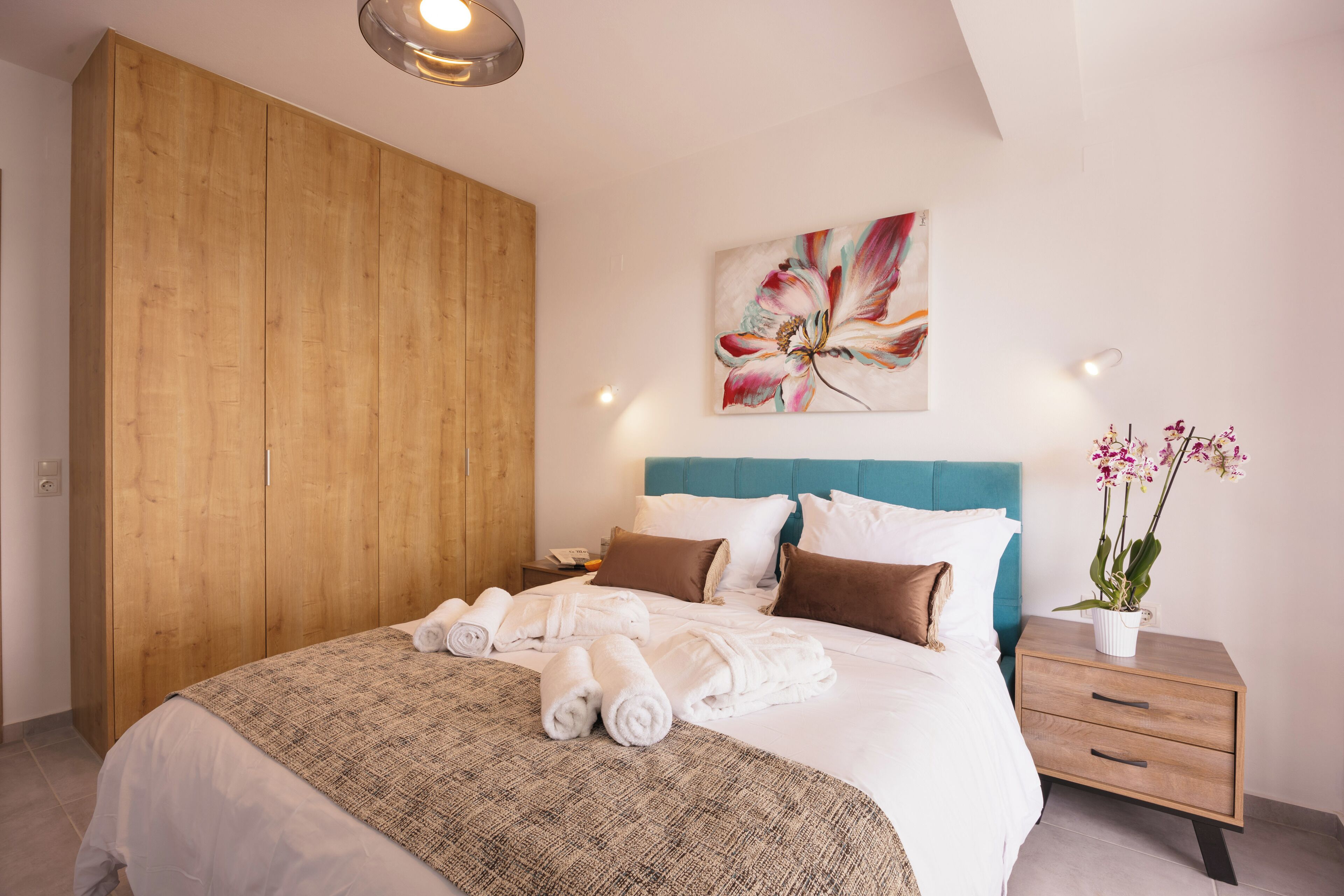 A neatly made bed with rolled towels, a wooden wardrobe, a teal headboard, a floral painting, a nightstand with a plant, and a wall socket.