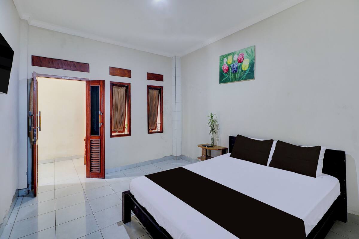 A bedroom with a large bed, a wooden nightstand, a potted plant, and a painting on the wall.