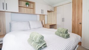 A bedroom with a large bed, striped bedding, two rolled towels, a wooden shelf with a plant, and a wardrobe.
