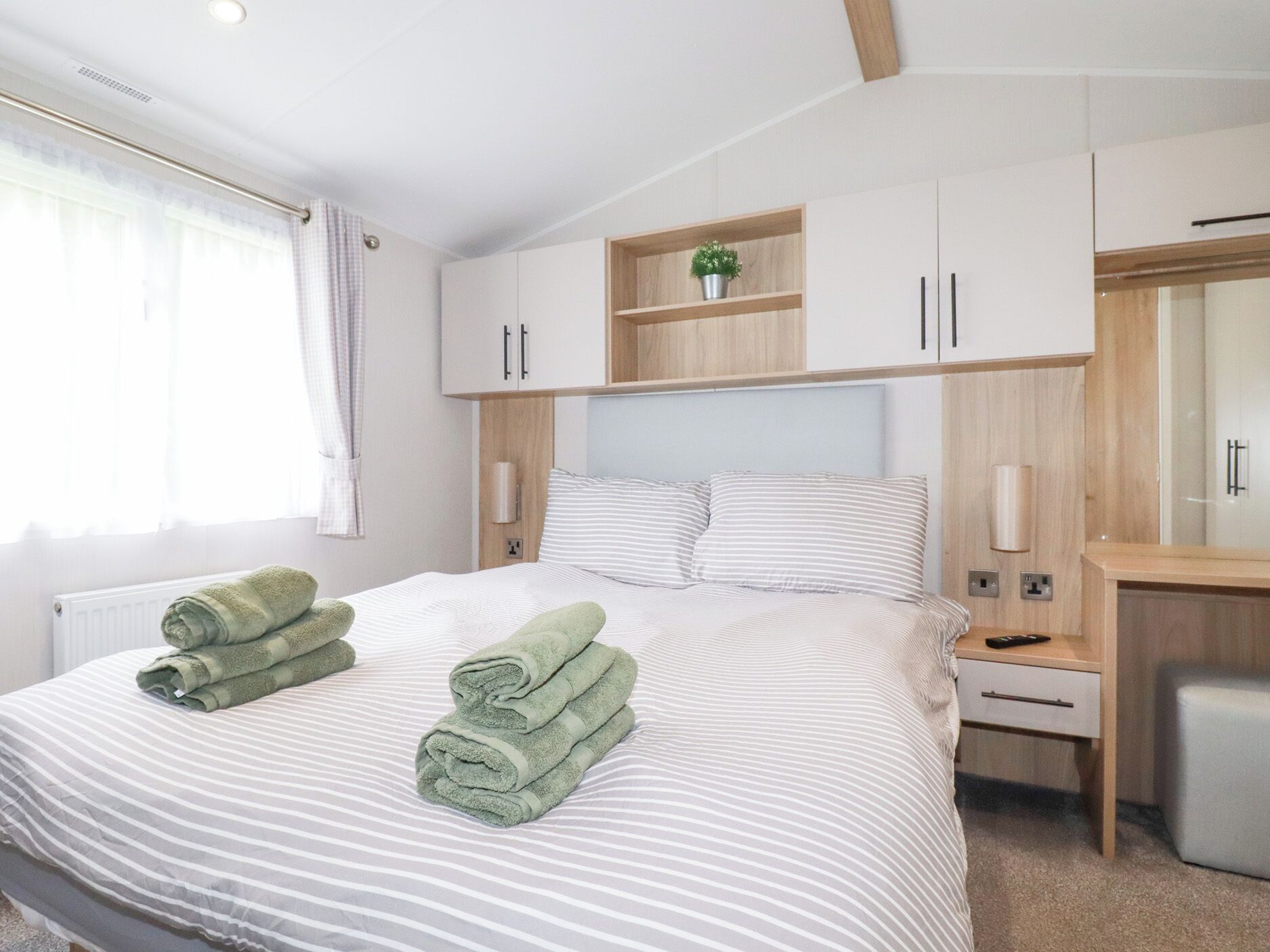 Een opgemaakt bed met gestreepte lakens en twee opgerolde handdoeken. Er is een houten hoofdbord, een nachtkastje met een lamp en een klein plankje met een plant.