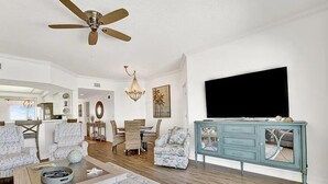 A spacious living room with a ceiling fan, a dining area with a chandelier, and a large TV on a teal console. - Coral Reef Unit #1304 (Beachfront 2 bedroom 2 bath condo on the 13th floor) (Panama City Beach)