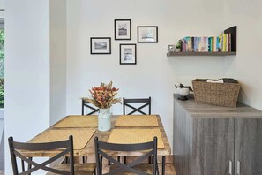 A dining area with a table, chairs, a vase with dried flowers, framed pictures on the wall, and a bookshelf with books.