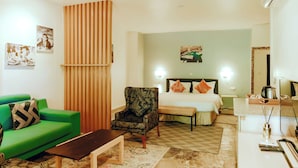 Une chambre d’hôtel avec un canapé vert, un fauteuil à motifs, un lit avec des coussins orange, une table de chevet avec une lampe et un grand miroir.
