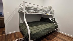 Una cama litera con una colcha verde y almohadas, un marco de metal blanco y un escritorio pequeño con una silla al fondo.