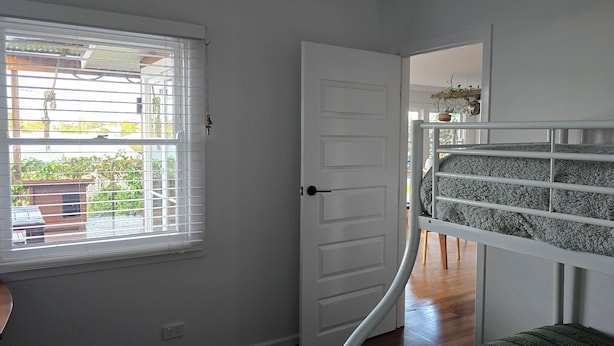 Una habitación con una cama litera, una ventana con persianas, una puerta y una planta pequeña sobre una estantería.