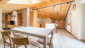 Une cuisine spacieuse avec des poutres en bois, un coin repas avec une table et des chaises blanches, et une horloge visible au mur.