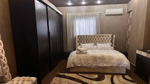 A bedroom with a large bed, a wardrobe, a chest of drawers, and a tufted ottoman.
