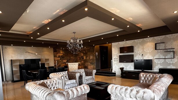 A spacious living room with a large chandelier, plush seating, a stone accent wall, and a flat-screen TV mounted on the wall.