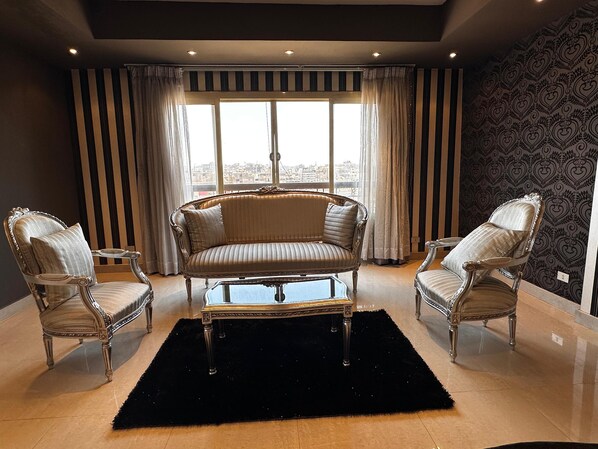 Ein geräumiges Wohnzimmer mit einer großen Couch, zwei Sesseln und einem Couchtisch. Das Zimmer verfügt über große Fenster mit Vorhängen, eine Wand mit Muster und einen schwarzen Teppich.