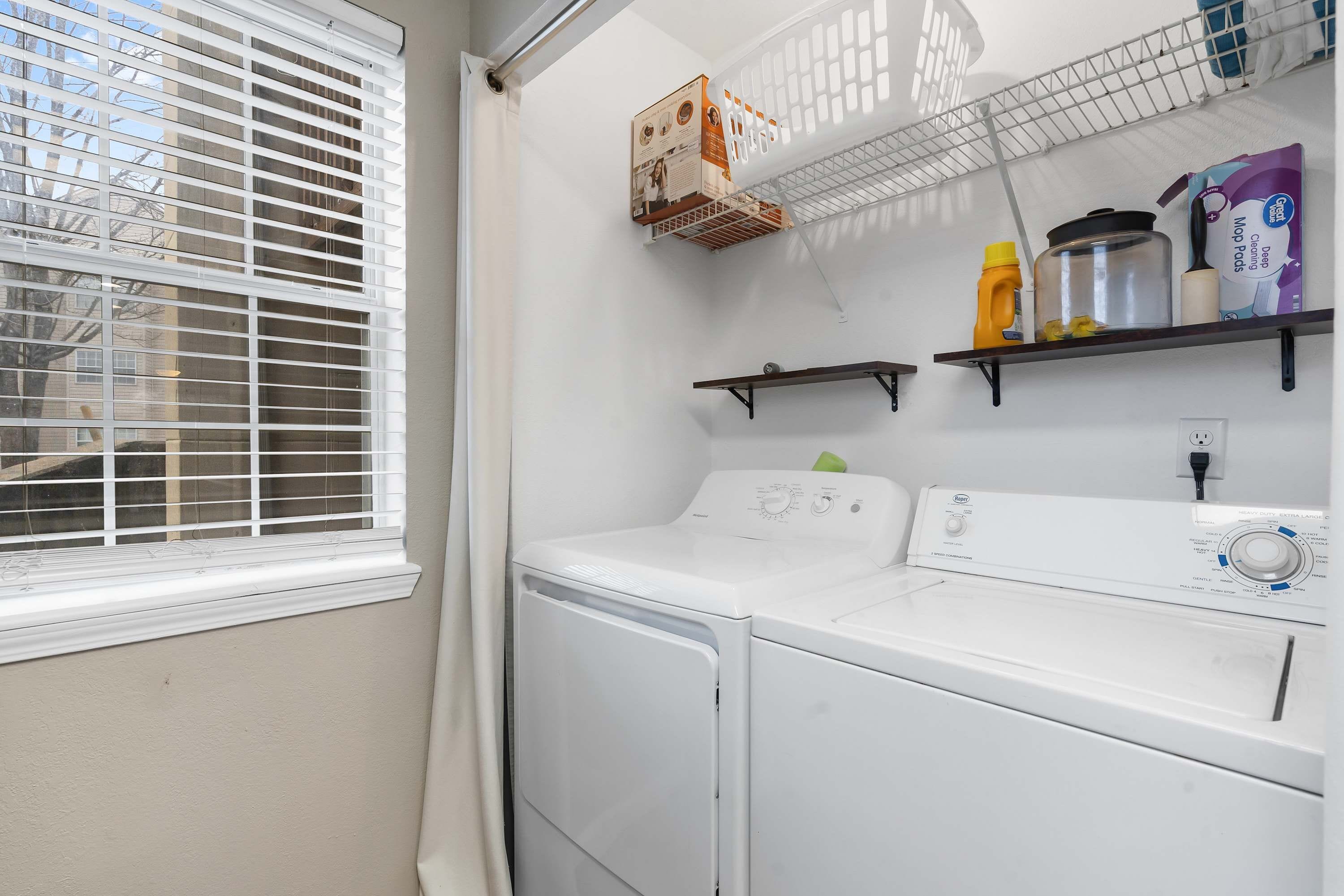 A laundry area with a front-loading washer and dryer, shelves with cleaning supplies, and a window with blinds.