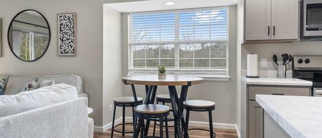 A modern kitchen with a dining area, featuring a round table with stools, a built-in oven, and a large window with blinds.