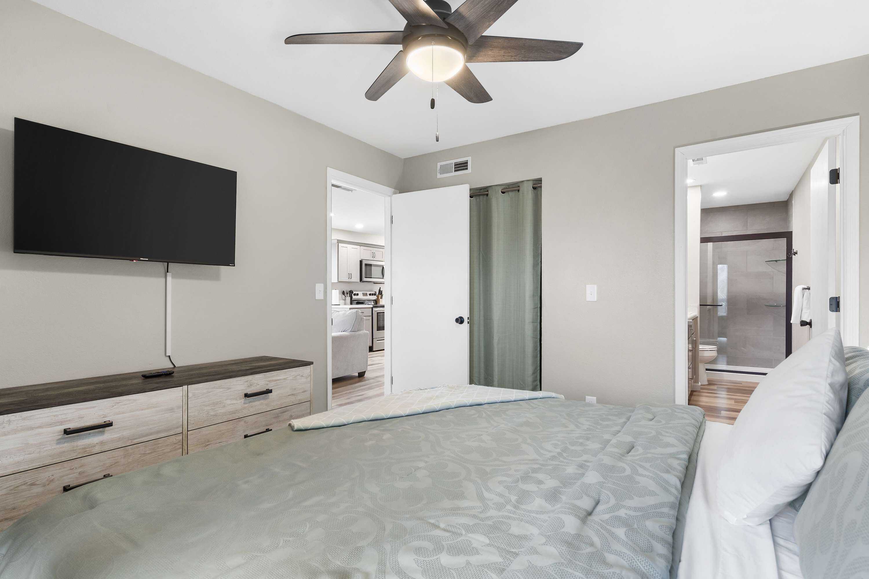 A bedroom with a bed, a TV, a ceiling fan, and a view into the kitchen.
