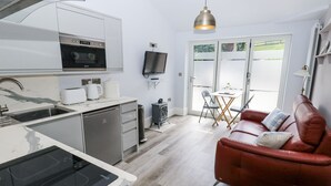 A modern kitchen with a built-in oven, microwave, and dishwasher. There is a dining area with a table and chairs, and a red leather sofa. A television is mounted on the wall, and a window provides a view of the outdoors. - Little Birchinlea (Glossop)