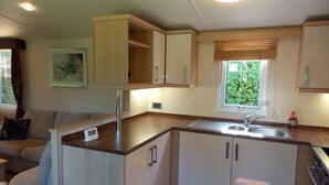 A compact kitchen with wooden cabinets, a stainless steel sink, and a window with blinds. - Emma 6 (Menai Bridge)