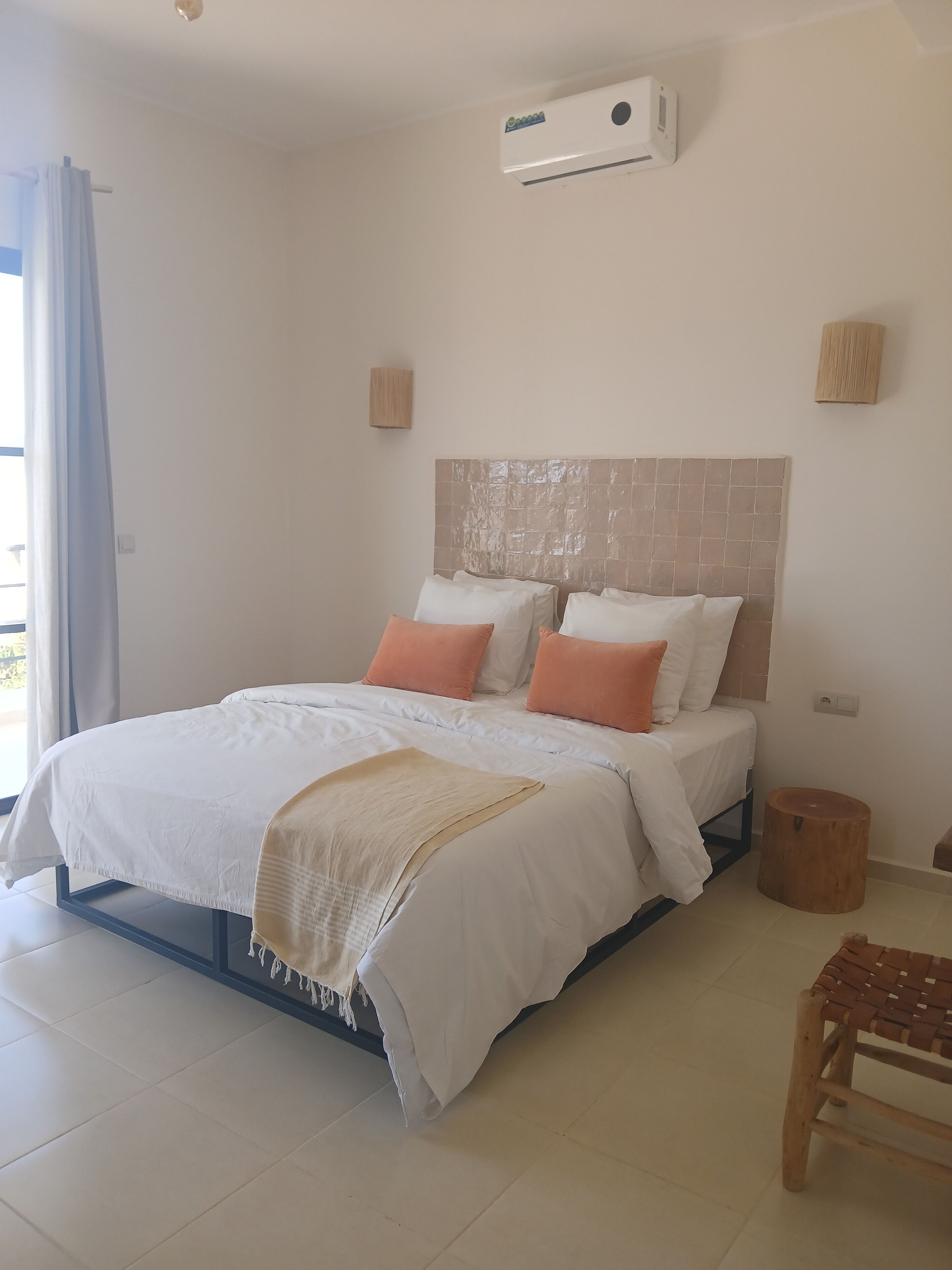 A bedroom with a bed, a bedside table, a wooden stool, and a wall-mounted air conditioner.