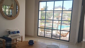 Un salón amplio con un gran ventanal, una mesa redonda de madera, un sofá con cojín y un espejo decorativo.