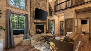 A spacious living room with a stone fireplace, wooden floors, a large sofa, and a TV mounted on the wall. - Cozy Cabin with Hot Tub, Fire Pit & Arcade Games (Broken Bow)