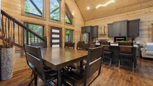 A spacious wooden cabin interior with a dining area, kitchen, and large windows. - Cozy Cabin with Hot Tub, Fire Pit & Arcade Games (Broken Bow)