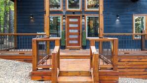 A wooden cabin with a front porch, a double door, and a small garden area. - Cozy Cabin with Hot Tub, Fire Pit & Arcade Games (Broken Bow)