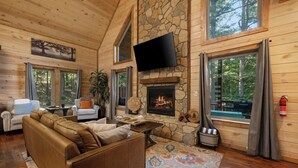 A log cabin interior with a stone fireplace, leather sofa, and large windows offering forest views. - Cozy Cabin with Hot Tub, Fire Pit & Arcade Games (Broken Bow)