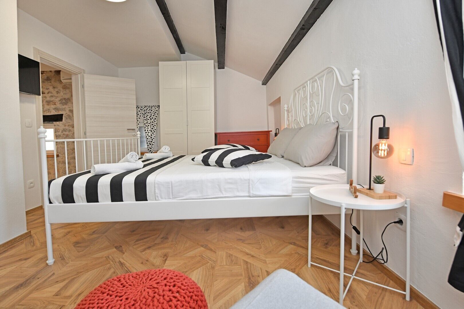 A modern bedroom with a white bed, striped bedding, a red knitted pouf, and a bedside table with a lamp.