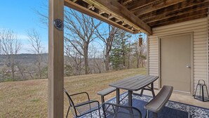 A covered patio with a table and chairs, a view of the surrounding landscape, and a door to the building. - Mossy Pines of the Ozarks (Branson)