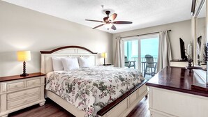 A bedroom with a bed, bedside tables, a ceiling fan, a window with curtains, and a view of the outdoors.