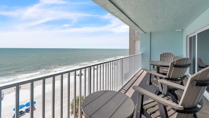 A balcony with a view of the beach and ocean. There are chairs and a table on the balcony.