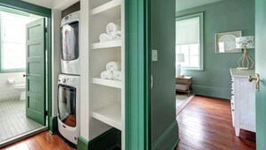 A laundry room with a washer and dryer, a shelf with folded towels, and a door leading to a room with a wooden floor and a window.