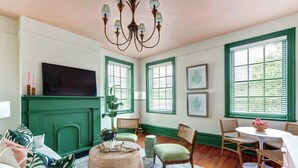 A cozy living room with a green fireplace, wicker furniture, and large windows with blinds.