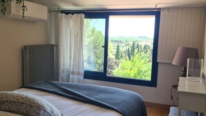 Un dormitorio con una ventana grande, un radiador, una mesita de noche con lámpara y una cama con manta.