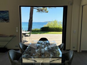 A dining area with a glass table and chairs, a view of the sea, and a wooden deck outside. - La Voile, Beachfront Villa (Grimaud)