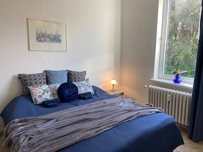 A bed with a blue quilt, multiple pillows, a bedside lamp, a window with a view of trees, and a radiator. - Apartment 'Dorfstraße' with Wi-Fi (Dänischenhagen)