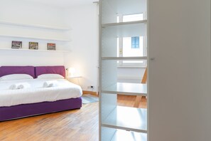 A modern bedroom with a purple bed, white walls, and a wooden floor. There are three framed pictures on the wall and a window with blue shutters. - Welcoming Flats by Lake Como (Como)