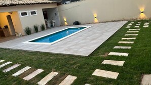 A backyard with a swimming pool, stone stepping stones, and a house with a tiled roof. - Cf07 Casa churrasqueira 5 min da praia (Cabo Frio)
