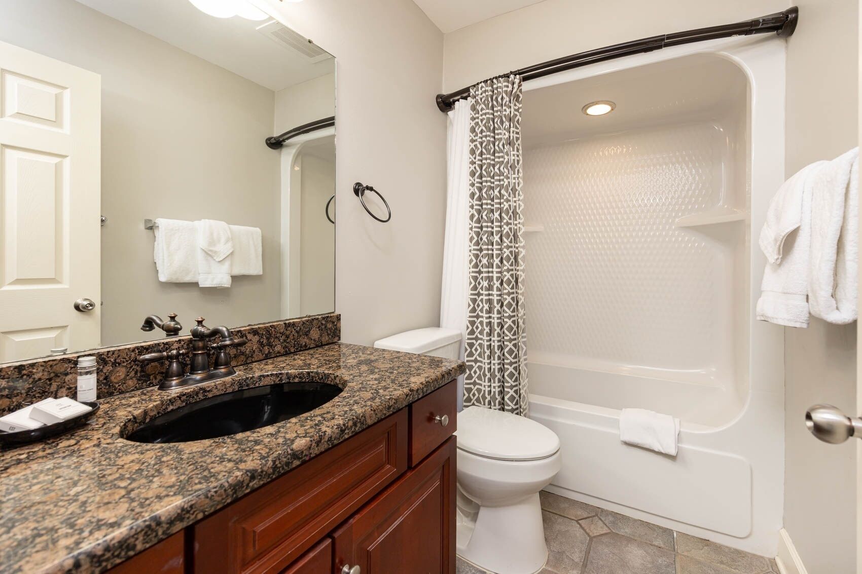 A bathroom with a granite countertop sink, a toilet, a bathtub with a shower curtain, and a mirror.