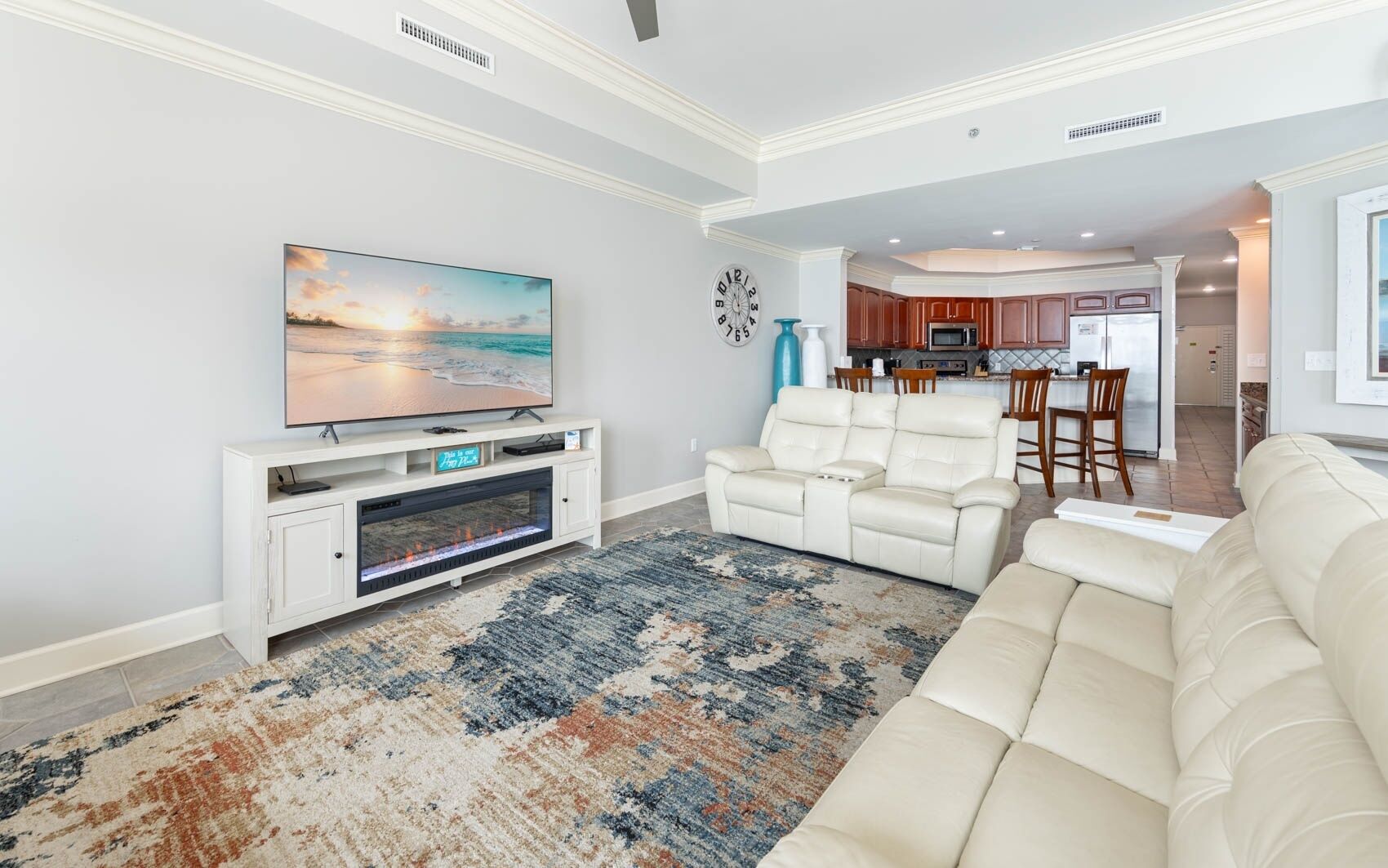 A living room with a fireplace, a large TV, a white sofa, and a kitchen in the background.